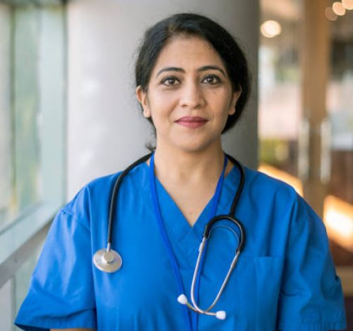 A female doctor or nurse of Indian descent wearing scrubs and a stethoscope poses next to a window in a hospital corridor and smiles confidently directly at the camera.